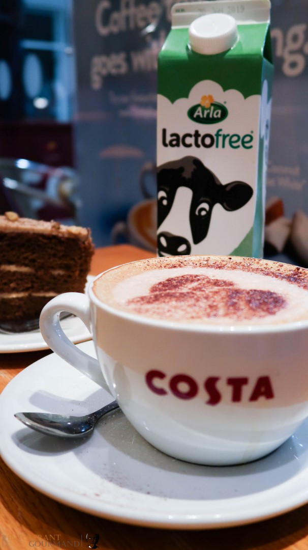 A large white cup of coffee made using Arla Lactofree milk on a table, with a carton of Arla lactofree milk in the background and available now at Costa Coffee! www.intolerantgourmand.com