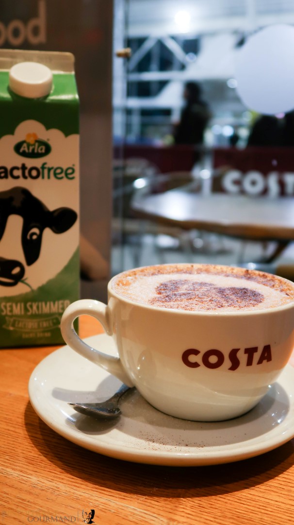 A large white cup of latte coffee made with Arla Lactofree milk on a table, with a carton of Arla Lactofree milk in the background, and available at Costa coffee now! www.intolerantgourmand.com