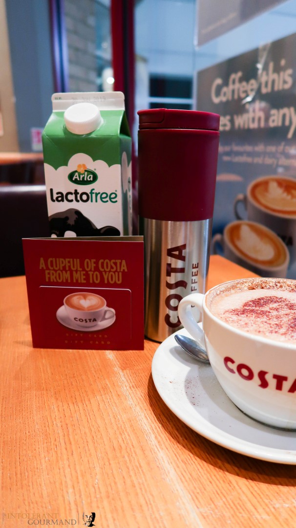 A large cup of latte coffee made with Arla Lactofree milk on a table, with a costa refill cup and costa voucher, along with a carton of Arla lactofree milk in the background. www.intolerantgourmand.com