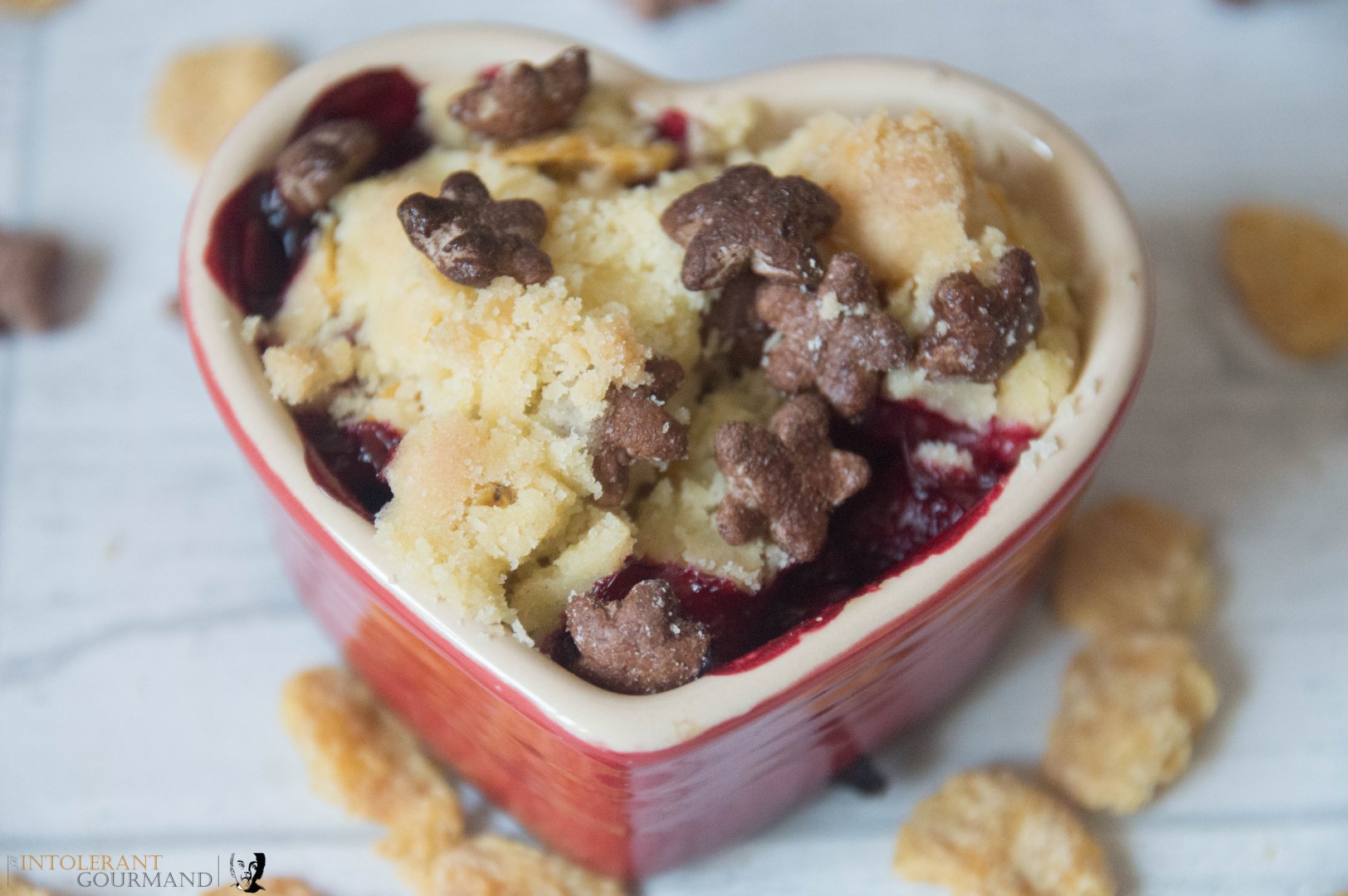 Mixed Berry Gluten Free Cobbler - a red heart shaped bowl filled with Mixed Berry Gluten Free Cobbler and a sprinkle of Cornflakes and Chocolate Stars on the white table. www.intolerantgourmand.com